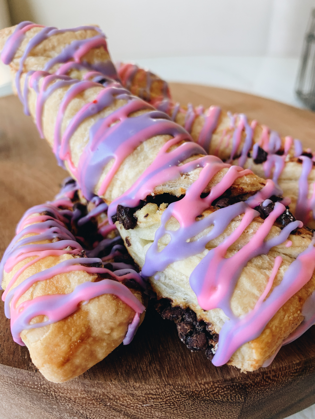 Three Cheshire Cat Tail Pastries stacked on top of each other, they are puff pastries with purple and pink icing.